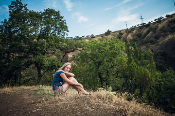 An attractive blonde girl in striped shorts and a blue t-shirt sitting along a path in the hills, around the trees, on a bright sunny day.