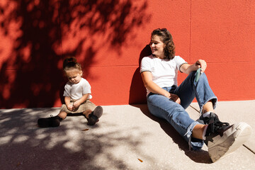 cheerful mother in jeans looking at toddler daughter while sitting near red wall.
