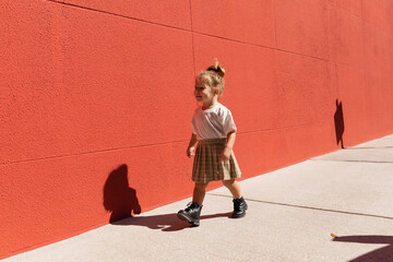 cheerful toddler girl in checkered skirt and white t-shirt walking near building with red wall.
