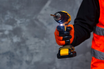 Male worker holds a close-up electric cordless screwdriver in his hands against the background of a construction tool and a concrete wall.