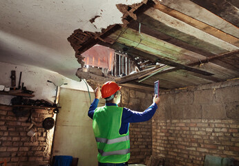 Building inspector. Man in a hard hat and a yellow reflective vest examines damaged structures and...