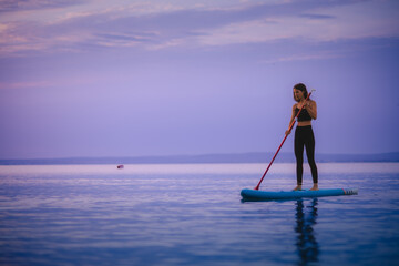 Young beautiful girl surfer paddling on surfboard on the lake at sunrise