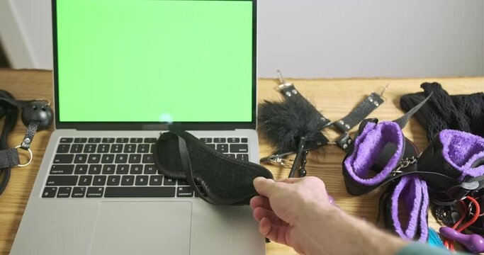 Man's hands put sex mask on clase on keyboard of laptop with green screen. Concept of online blind dating, sex shop, erotic bdsm games.