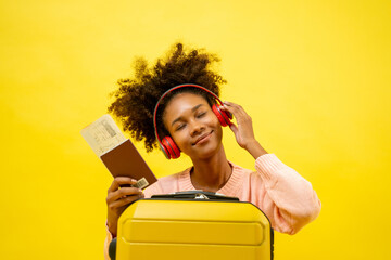 Portrait of young attractive traveling african american woman curly hair with headphone, baggage, passport and boarding pass in studio on yellow background.