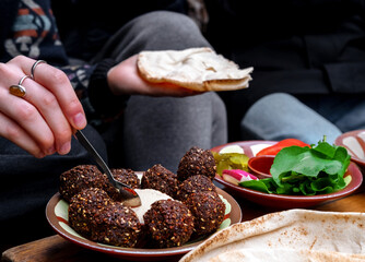 Close-up of women's hands holding bread and spreading hummus. A plate of falafel and vegetables on the table. Middle East cuisine. - obrazy, fototapety, plakaty