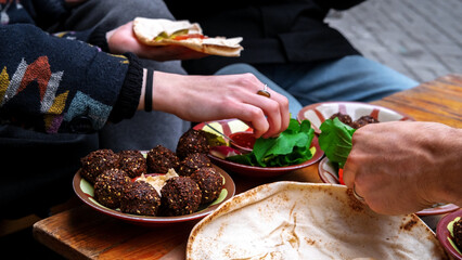 Close-up of female and male hands holding bread and spreading hummus. A plate of falafel and...