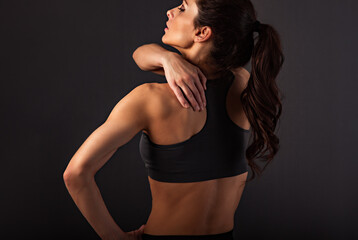 Fototapeta na wymiar Female sporty muscular with ponytail doing stretching workout of the shoulders, blades in sport bra, holing the neck the hand on dark grey background with empty space.