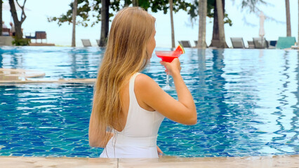Woman relaxing in infinity pool at luxurious hotel, take glass with tropical cocktail, drinking refreshment drink. Concept of a girl enjoying vacation in sea beachfront resort in heat - obrazy, fototapety, plakaty