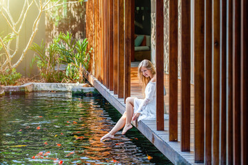 Woman relax in luxury spa with fish pond, enjoy bright morning and nature, relaxing and having good time sitting on wooden bridge barefoot. female playing feet in water at outdoor pond. Slow motion