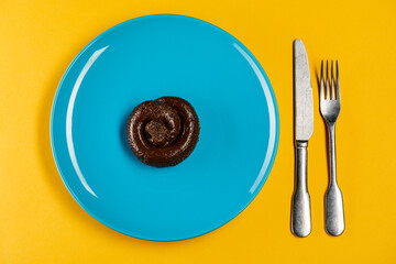 Plastic excrement lies on a plate between a fork and a knife close-up. The concept of an inept cook, fake news, shitty restaurant, bad food, taste of shit