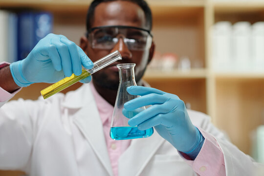 Selective focus on gloved hands of clinician pouring liquid from flask into beaker while carrying out scientific experiment in laboratory