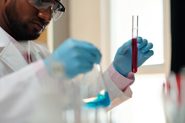 Focus on gloved hand of chemist holding flask with crimson liquid substance before pouring it into beaker during experiment