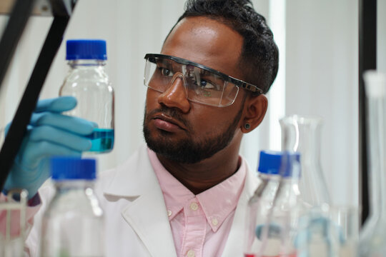 Serious young male clinician looking at glassware containing blue liquid while carrying out scientific experiment in laboratory