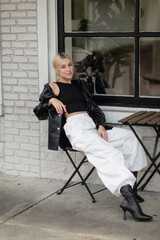 Fototapeta na wymiar full length of blonde woman in leather shirt jacket and cargo pants sitting in outdoor cafe on street in Miami.