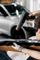 A worker covers a car spoiler with a film in the workshop using a hair dryer and a spatula close-up...