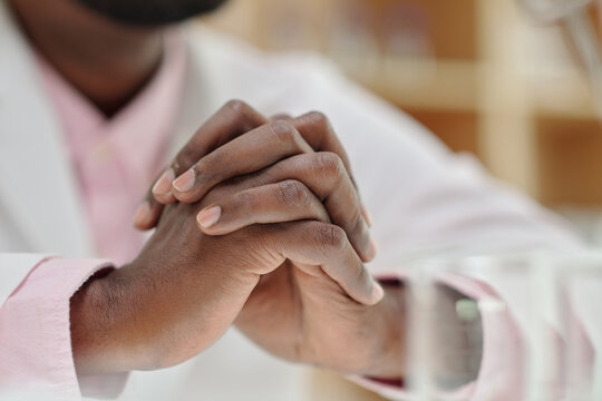 Close-up of clasped hands of young African American male clinician wearing lab coat concentrating while thinking during work