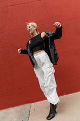 full length of cheerful woman in cargo pants and leather shirt jacket standing near red wall in...