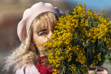 A young woman stands with a bouquet of yellow mimosa and sniffs flowers in the park. The concept of the Spring holiday - March 8, Easter, Women's Day