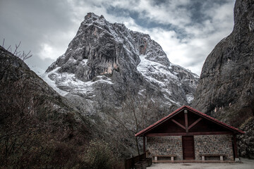 Fototapeta na wymiar A small building is situated against a mountain range, surrounded by tall trees and a cloudy sky