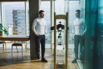 A young handsome man in a white sweater is standing in a spacious bright office. The concept of a modern successful person. Young smiling guy in open office space