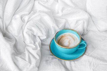 A blue cup with cappuccino stands on the bed next to the blanket