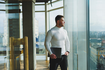 Fototapeta premium A young man in a modern workspace stands near a large window. A man in casual clothes with a laptop in his hands. Remote work. Nice young manager