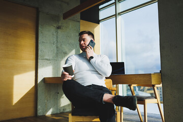 Cute young male freelancer drinking coffee while sitting in bright workspace working remotely. A man in a white sweater and trousers works happily in the office