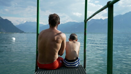 Father and son sitting by lake pier together overlooking Swiss mountains at Lake Geneva. parent and child bonding. Family enjoying summer vacations