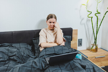 Young European beautiful woman sitting on bed with coffee, working with computer at home, remote work. Woman happy and smiling in bed at home.