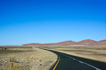 A long asphalt road in the desert in perspective against a blue sky. A jeep drives in the distance along the road.