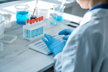 A female researcher sits at a workplace in a laboratory, behind a personal computer monitor. Against the background research statistics. Pharmaceutical medical worker in protection works at keyboard.