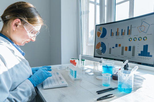 A female researcher sits at a workplace in a laboratory, behind a personal computer monitor. Against the background research statistics. Pharmaceutical medical worker in protection works at keyboard.