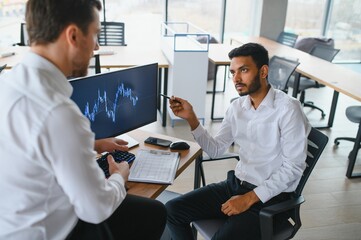 Two successful trader sitting in office and looking at screen, checking cryptocurrency information data on finance market graph, pointing on monitor