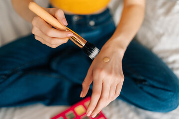 Close-up cropped shot of unrecognizable female put makeup foundation on hand and going to use brush to apply makeup to face sitting on bed in bedroom. Concept steps of make-up applying.