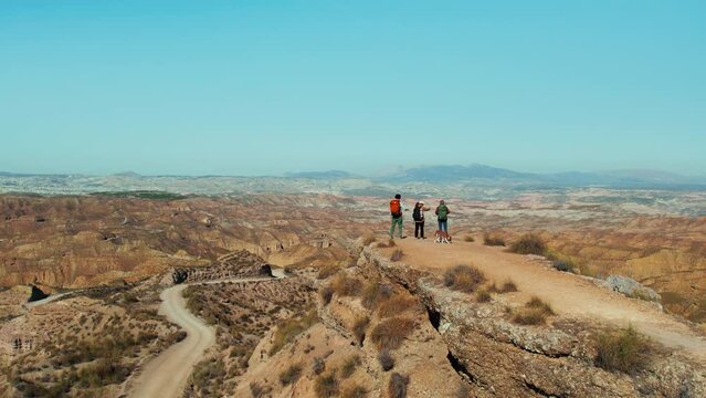 Group of hikers with dog embarks on an outdoor journey up a desert hill, seeking adventure and scenic nature. At the edge of the hill, tourists revel in the peaceful view of surrounding mountains. 