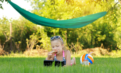 kid girl is eating piece of pizza and playing on laptop lying on grass on summer sunny day. the child has lunch after active games. hammock and ball on the background.