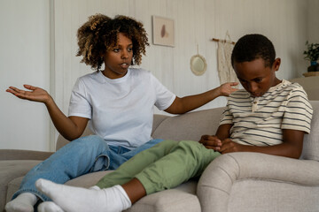 Dissatisfied African American woman mother scolding upset son for bad behavior while sitting together on sofa in living room. Disappointed parent mom talking with kid, disciplining child at home