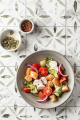 Traditional italian tomato salad panzanella with mozzarella, capers, red onion, croutons, cucumbers and basil. Summer salad on printed tile background with copy space, top view