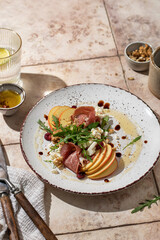 Salad of arugula and prosciutto or ham, sliced apple, cheese, walnuts on rustic plate in composition with cutlery and water glass on pink tile background in hursh light. Copy space