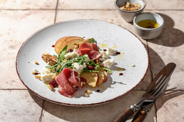 Salad of arugula and prosciutto or ham, sliced apple, cheese, walnuts on rustic plate in composition with cutlery on pink tile background in hursh light
