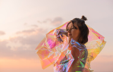 Young woman posing with holographic foil against sunset sky. Dreamy self expression concept fashion...