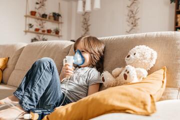 Little boy makes inhalation with a nebulizer at home lying on the couch. Cough prevention and...