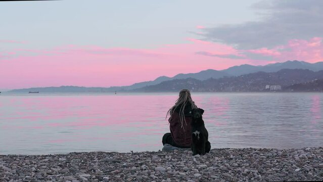 Woman sitting and meditating on the seashore during dawn. The dog is waiting nearby. Beautiful pink sky mountains on the horizon and a dolphin in the sea