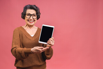 Portrait of senior aged mature woman with tablet computer, grandmother isolated on pink background....