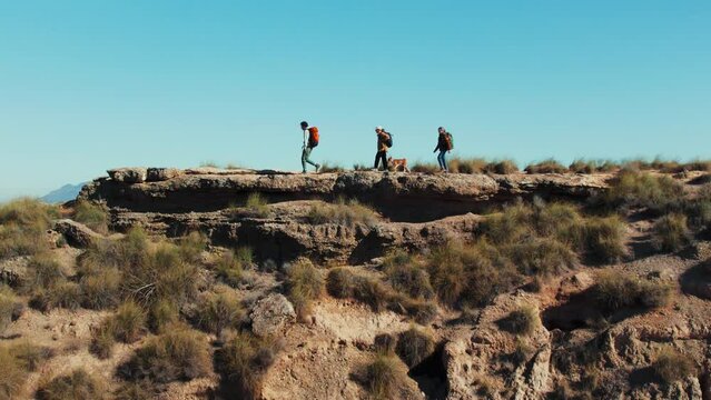 Touristic group of hikers with dog and backpacks reaches top of desert hill, taking in the scenic views of the mountains. The adventure-filled journey offers a peaceful escape into nature's beauty.