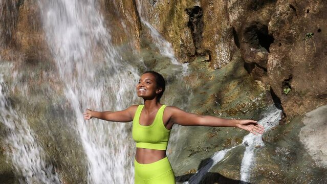Young Black Woman Standing Outdoors In Front Of Waterfall Arms Wide Open. Practicing Mindfulness