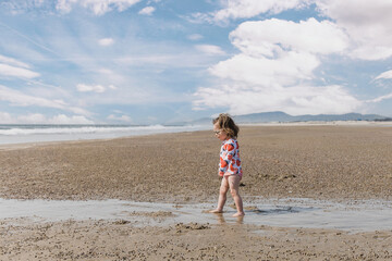A small toddler girl walking on the sandy ocean beach in a swimsuit and sunglasses