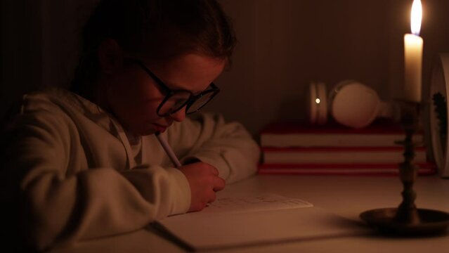 Schoolgirl distance learning at home in a complete darkness without electric lights. Teen kid uses candlelight to do her school homework during blackout. Energy crisis concept by. Blackout in Ukraine.