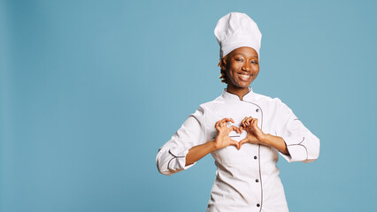 Female cook showing heart shaped symbol on camera, expressing romantic gesture and wearing cooking apron. Young adult chef being flirty with romance love sign, culinary industry. - obrazy, fototapety, plakaty