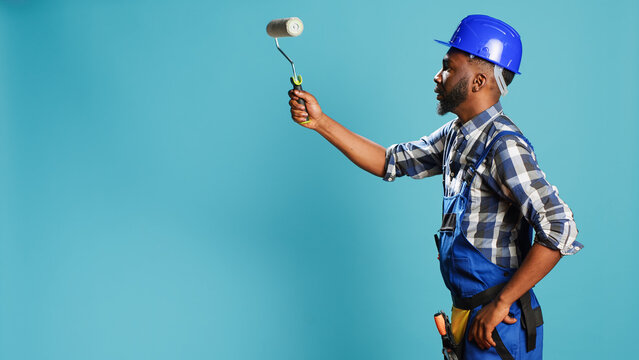 Industrial worker in uniform painting walls with roller, using renovation instrument over blue backdrop. Professional craftsman with hardhat working with rolling paint brush to change color.